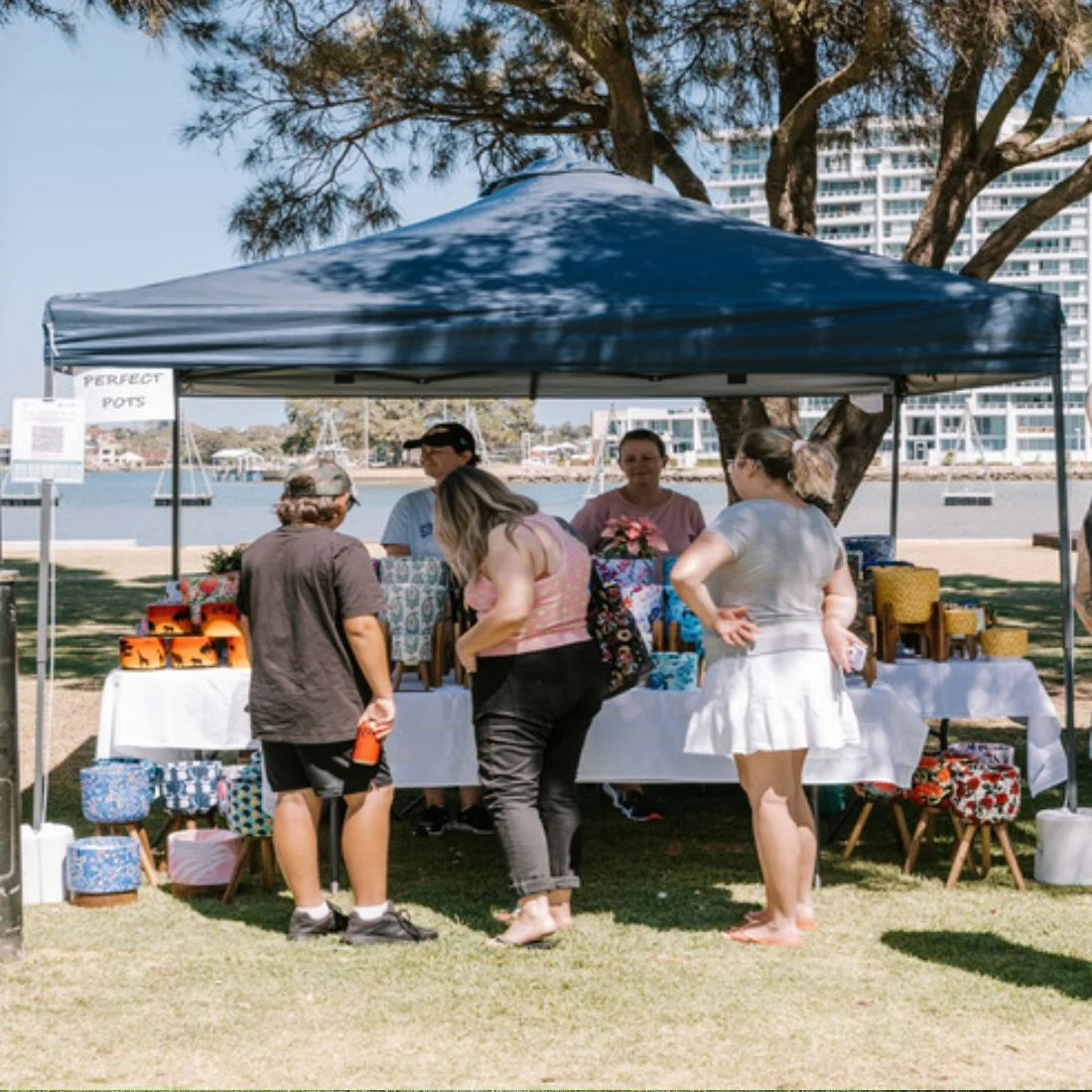 People browse colorful pots and planters displayed under a dark blue canopy at an outdoor market near a waterfront, with trees and apartment buildings in the background.