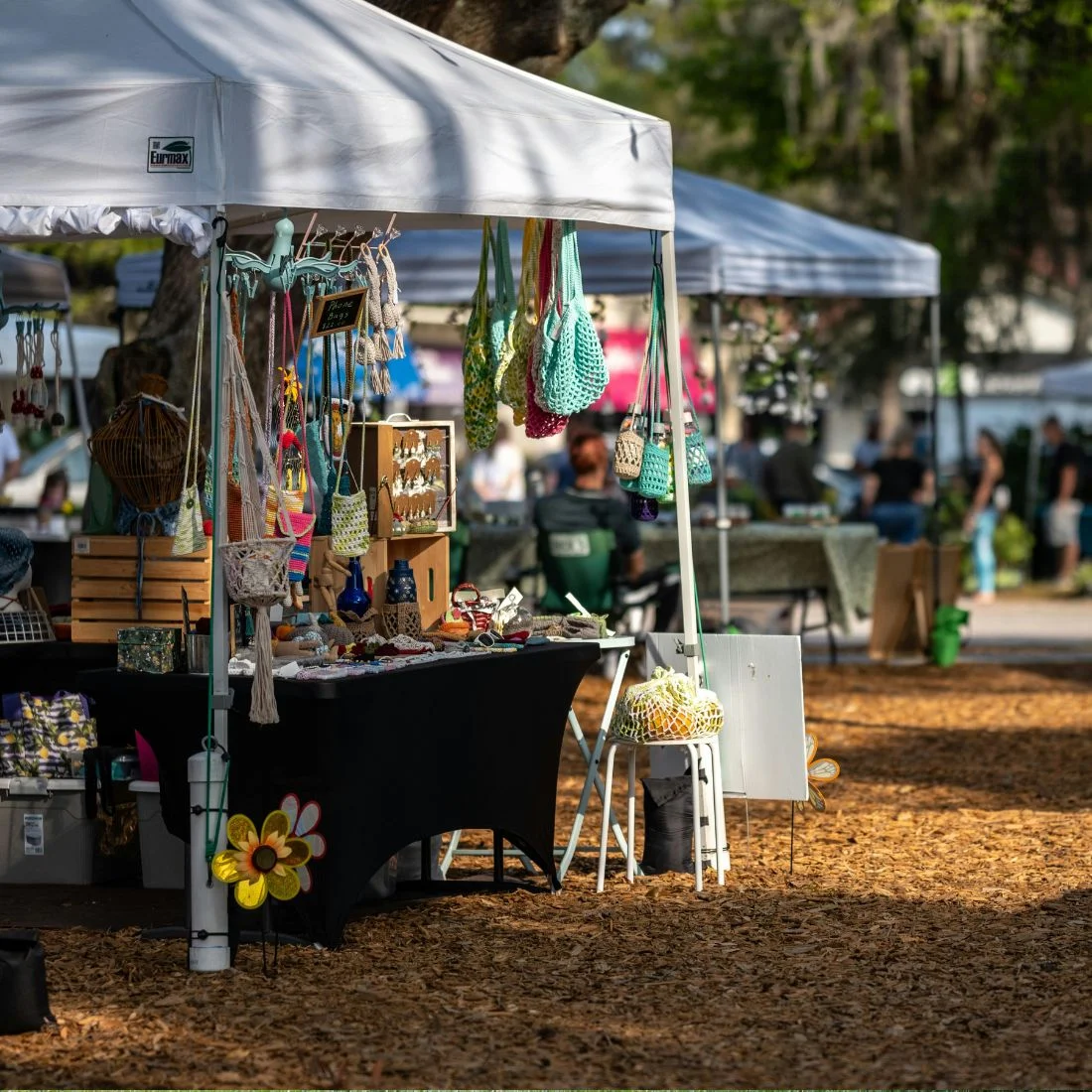 Outdoor market stall under a white canopy selling handmade crafts and bags, with people and other booths visible in the background.