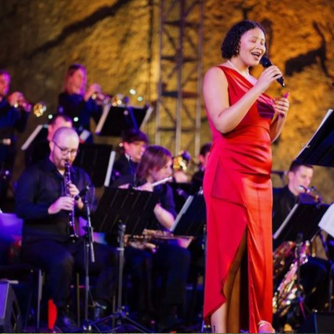 A woman in a red dress sings into a microphone on stage, accompanied by a seated band of musicians playing various instruments in the background. The setting is warmly lit with a golden-yellow backdrop.