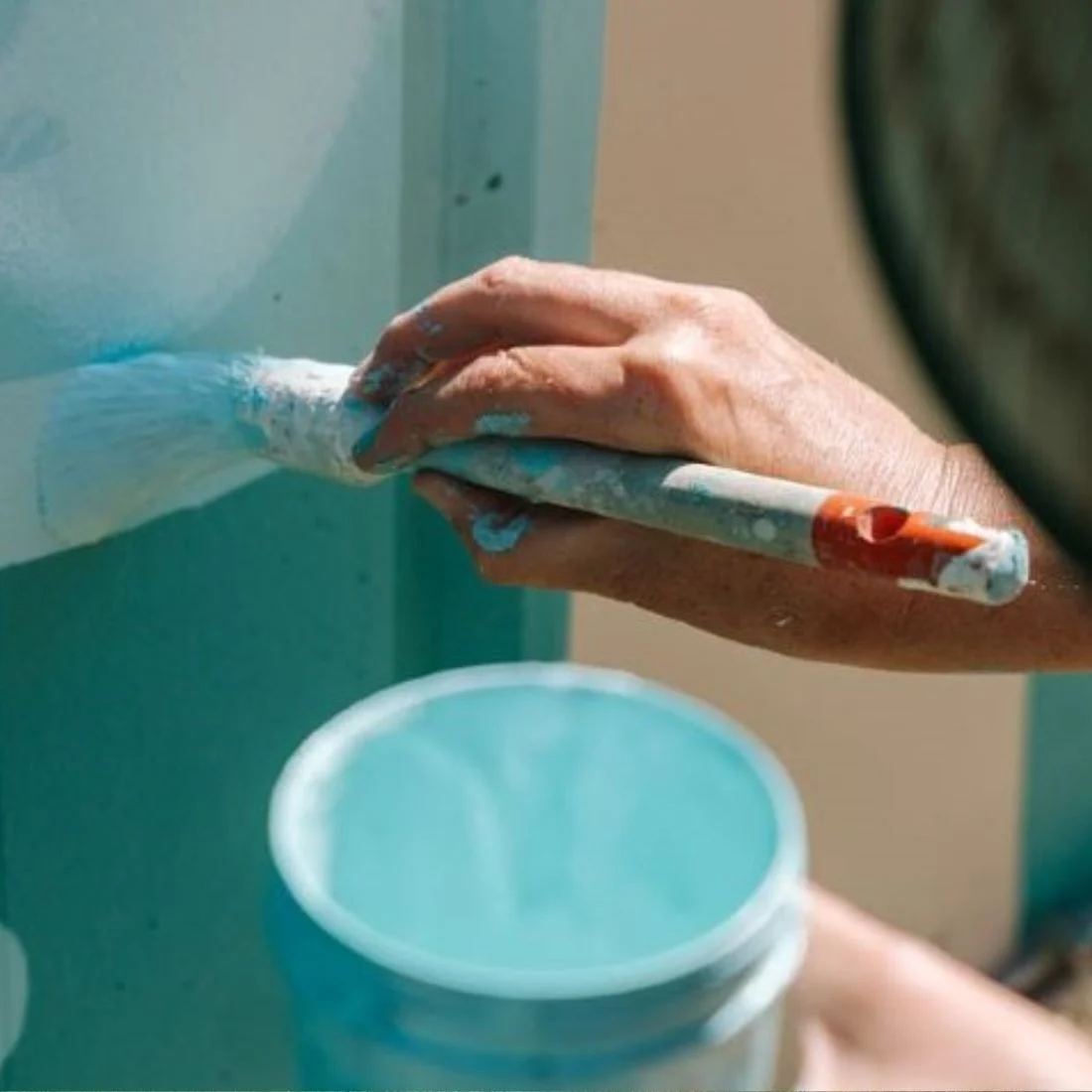 A close-up of a hand holding a paintbrush, painting a surface light blue. An open container of blue paint is nearby, and the painter’s fingers are speckled with paint.