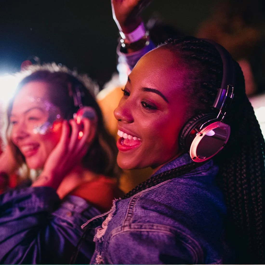 Two women smiling and dancing while wearing headphones at a night event, with colorful lights illuminating their faces.