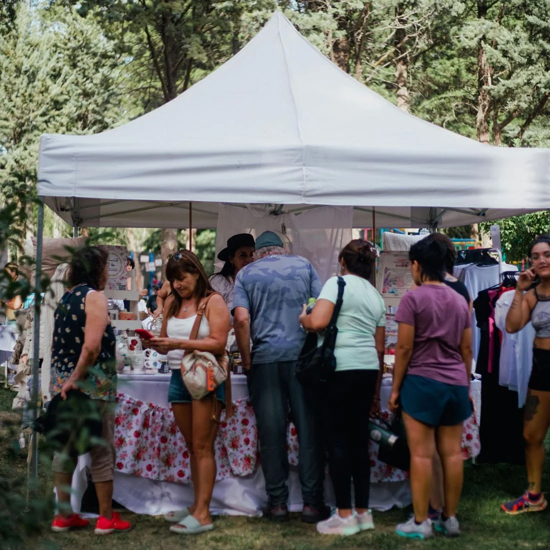 A group of people browse and shop at an outdoor market stall under a white canopy, surrounded by trees and greenery on a sunny day. Some people are looking at items on display, while others wait or chat nearby.