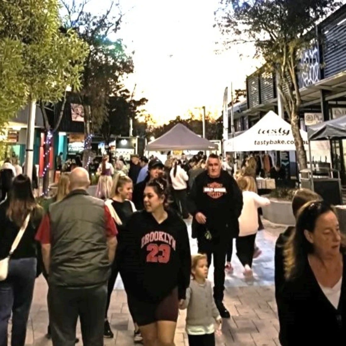 A busy outdoor market scene at sunset, with people walking along a pedestrian street lined with trees, market stalls, and shops. Some are shopping, while others are socializing.