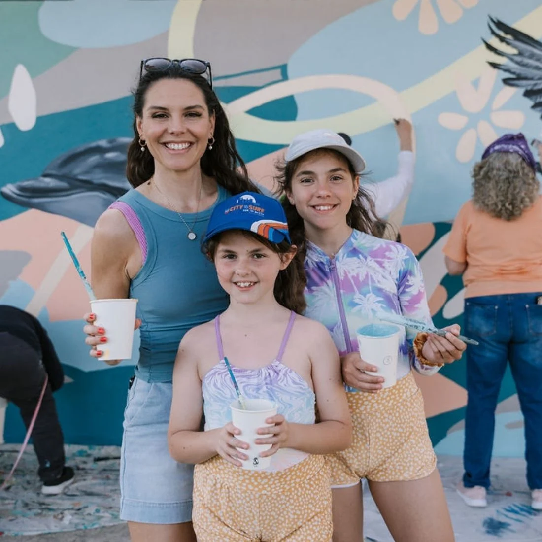 A woman and two girls smile while holding drinks in front of a colorful mural. They wear casual summer clothes and hats. Other people are painting the mural in the background.