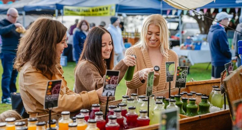 Three women smile and sample colorful bottled juices at an outdoor market stall, with more people and vendor tents visible in the background.