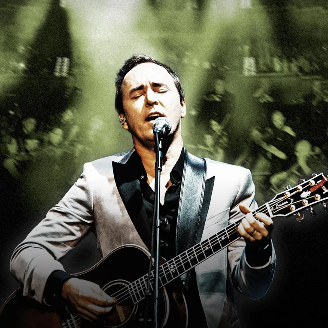 A man in a silver jacket sings passionately into a microphone while playing an acoustic guitar on stage, with green-yellow lights and a blurred audience in the background.