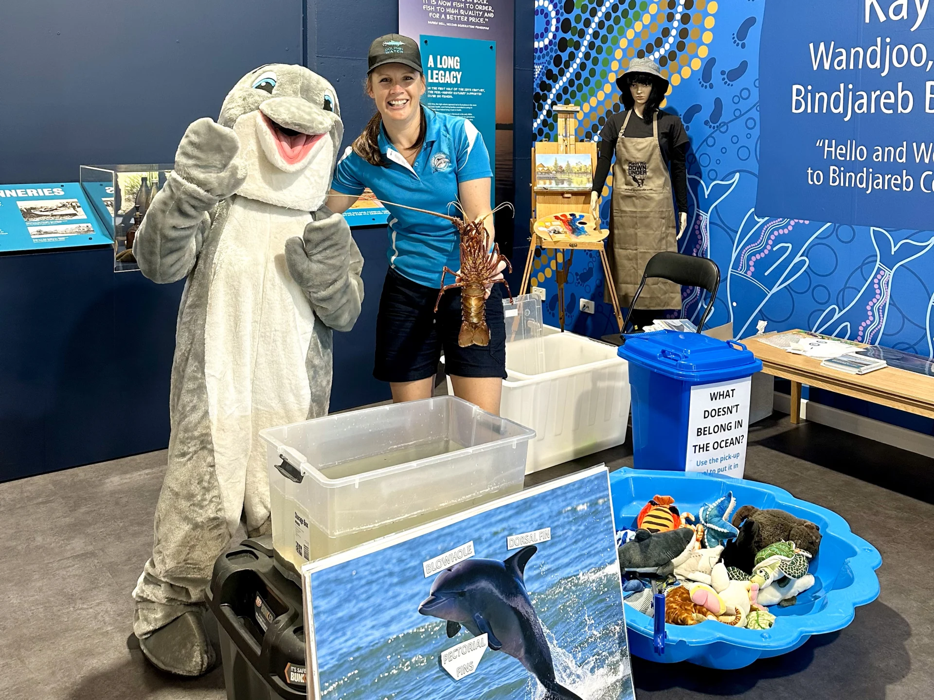 A woman in a blue shirt and cap stands next to a smiling dolphin mascot, holding a lobster. They are at an educational marine exhibit with posters, bins, props, and display tables about ocean conservation.