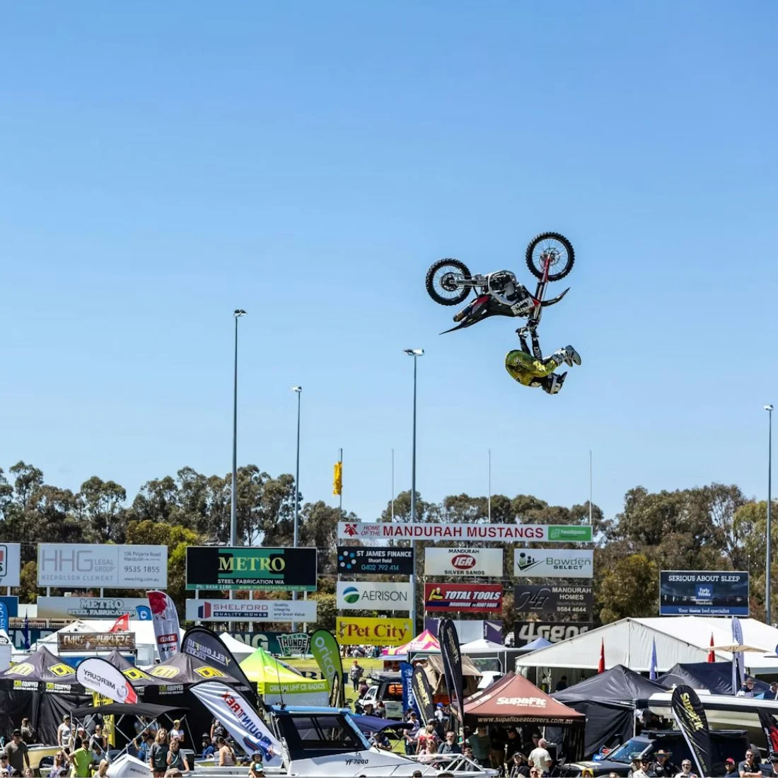 A motocross rider performs a mid-air backflip on a motorcycle above a crowd and tents at an outdoor event under a clear blue sky.