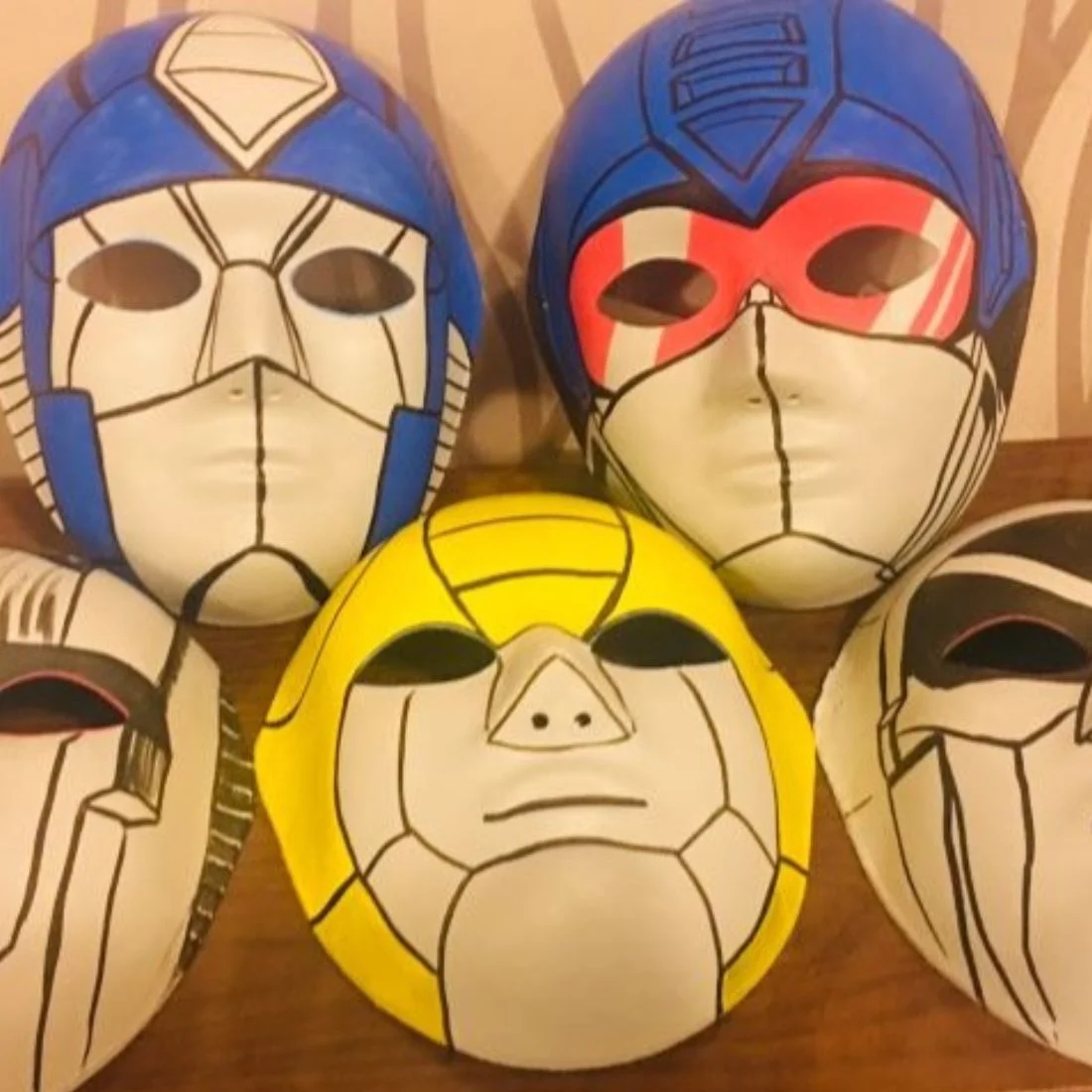 Six masks inspired by Transformers characters, each with robotic designs and bold outlines. Three are white with blue, red, or black accents, one is bright yellow, and two have black details. They are arranged on a wooden surface.