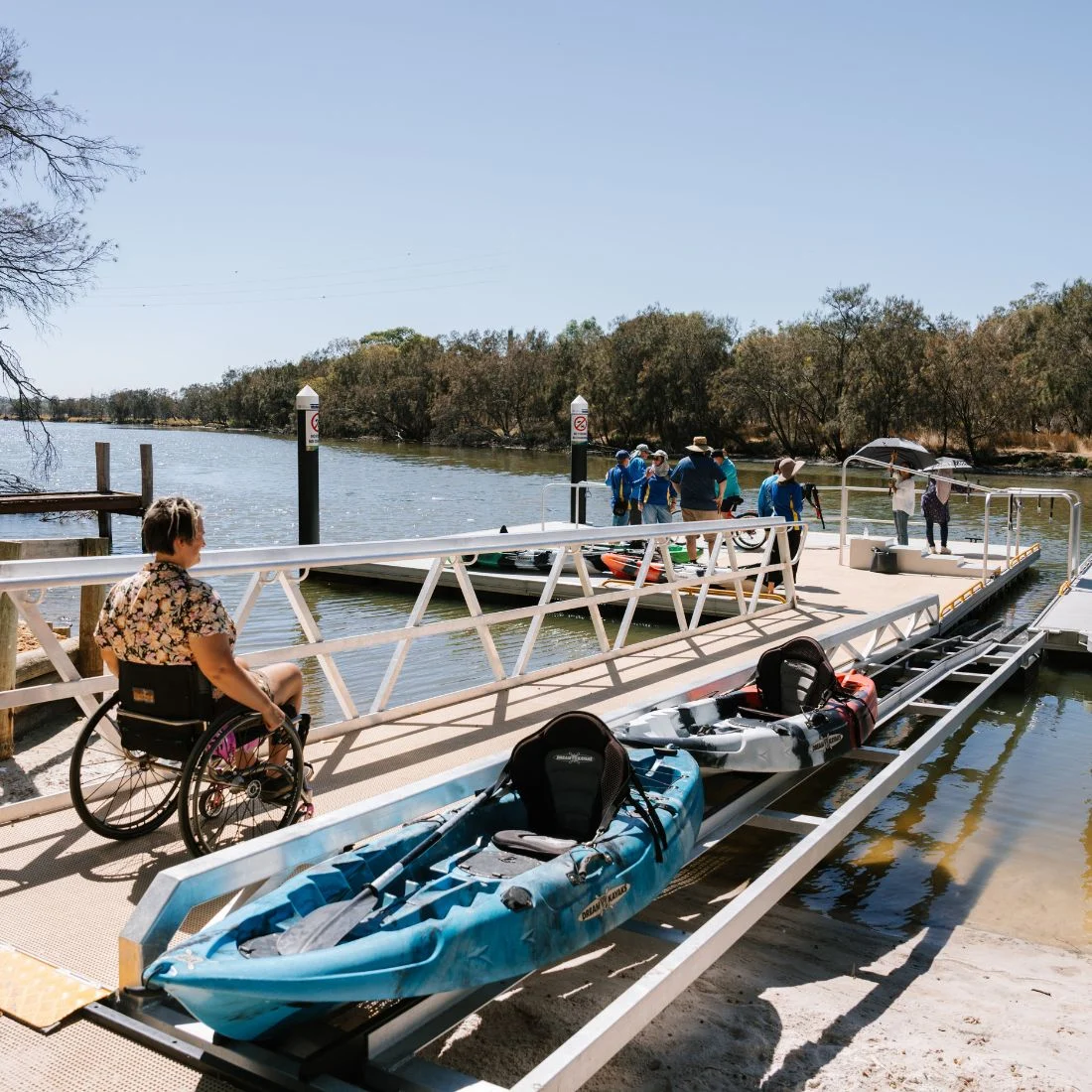 A person in a wheelchair approaches a ramp leading to a dock by the water, where kayaks are positioned and a group of people are gathered in the background under clear skies.