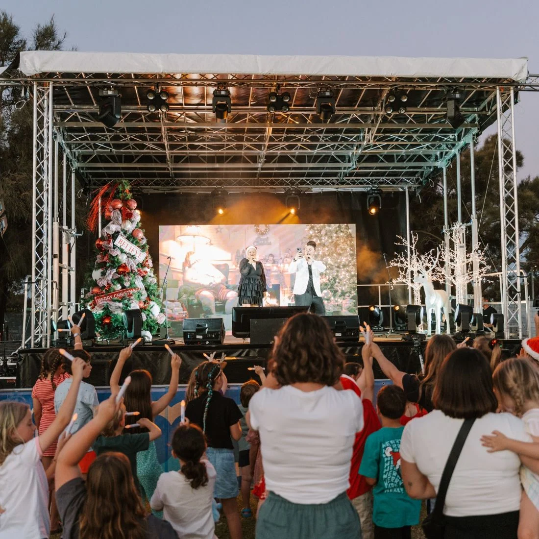 Two singers perform on a stage with a Christmas tree and lit reindeer, while a crowd of children holds glowing light sticks toward them.