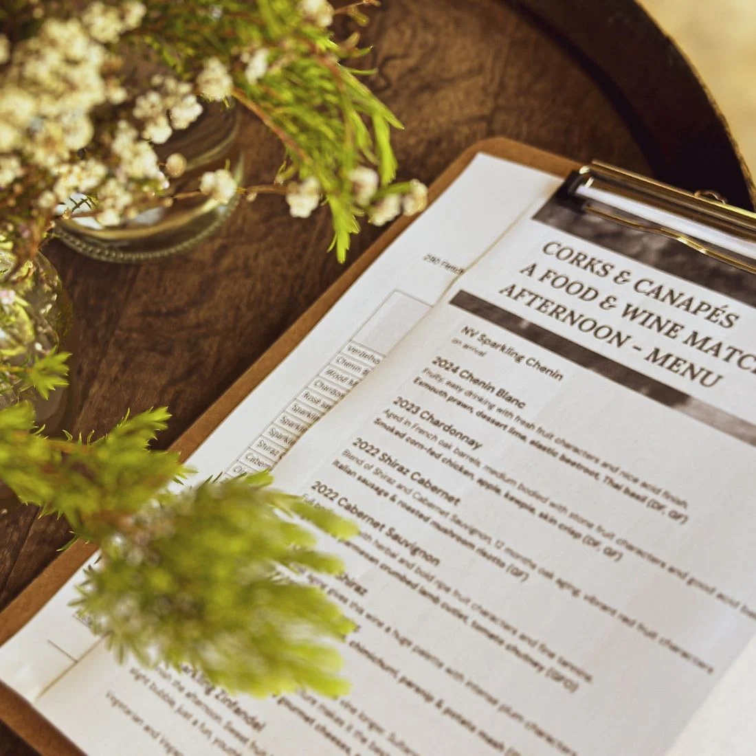 A clipboard with a printed menu titled Corks & Canapés: A Food & Wine Matching Afternoon rests on a wooden table beside a vase with green leaves and white flowers