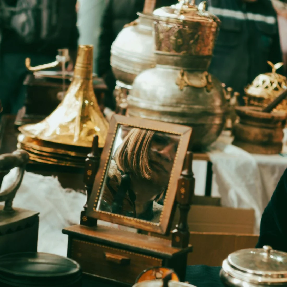 A small mirror on a table at an antique market reflects the head of a person with short, light brown hair. Surrounding the mirror are various brass and metal vessels and objects.