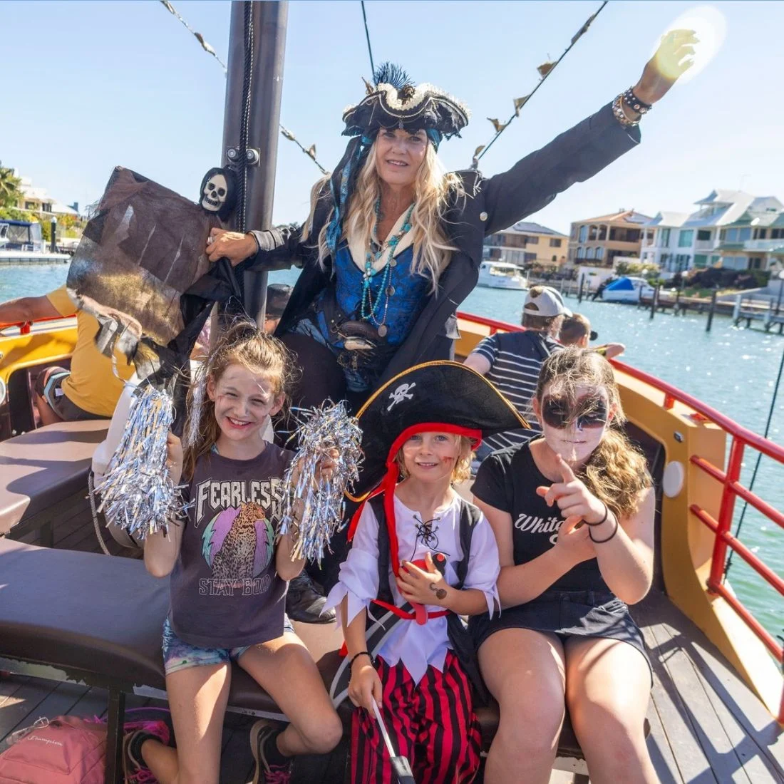 Four children in pirate-themed costumes and face paint pose and smile on a boat with an adult dressed as a pirate behind them. Water, houses, and a bright sky are visible in the background.