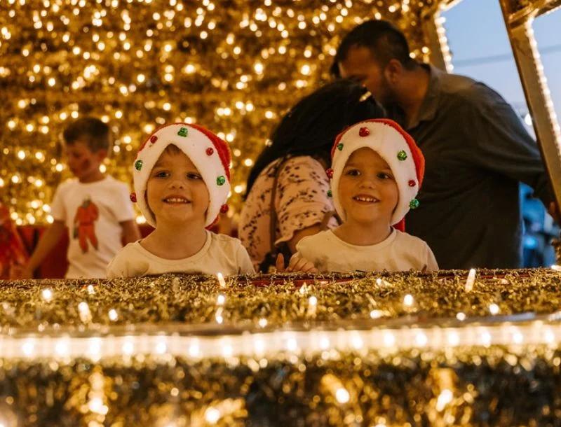 Two young children wearing festive Santa hats smile in front of a display decorated with gold tinsel and glowing holiday lights. Several adults and another child are blurred in the background.