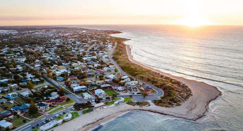 Aerial view of a coastal town at sunset, showing houses near the shoreline, roads, and a sandy beach with waves gently approaching the land. The sun is low on the horizon over the ocean.