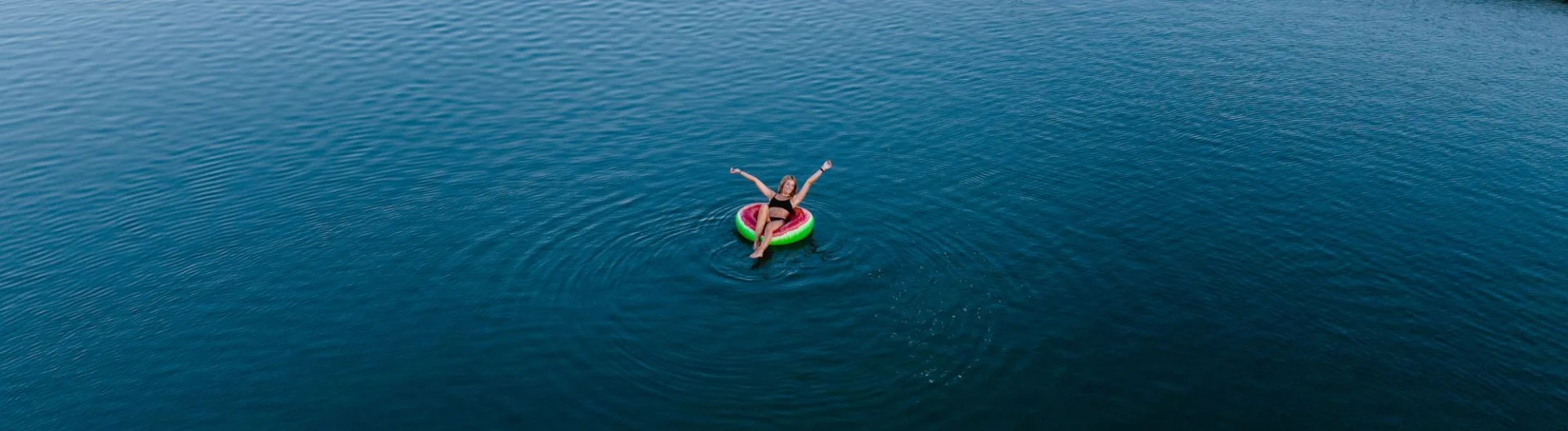 A person relaxes on a colorful inflatable ring in the middle of a calm, blue body of water, stretching their arms and legs out. Gentle ripples spread out around them.
