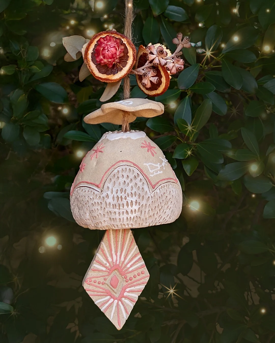 A decorative ceramic Christmas ornament with etched patterns hangs by a string, adorned with dried orange slices and flowers, against a leafy green background with glowing light effects.