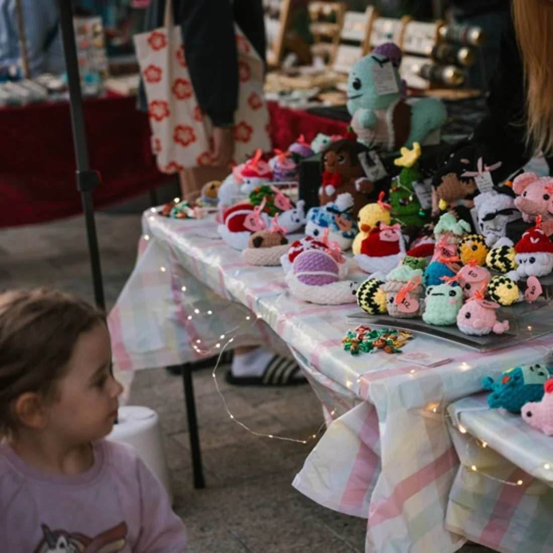 A young child looks at a market table covered with handmade crochet plush toys and colorful trinkets, with fairy lights draped along the edge of the table. Other market stalls are visible in the background.