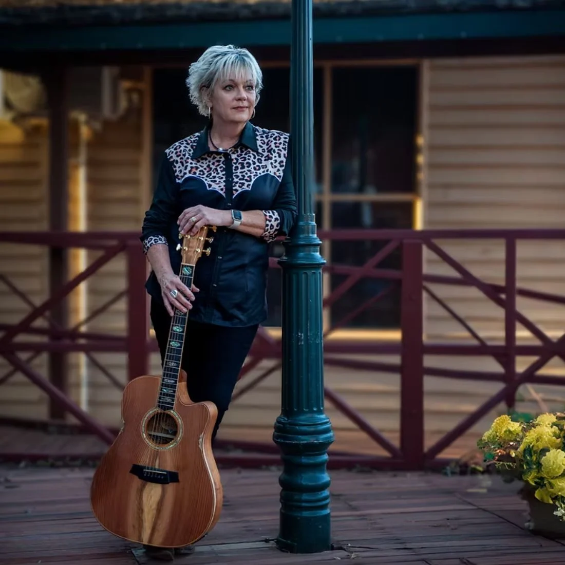 A woman with short gray hair, wearing a black shirt with a leopard print pattern, leans against a lamppost on a wooden deck and holds an acoustic guitar. There is a wooden building and a pot of yellow flowers in the background.