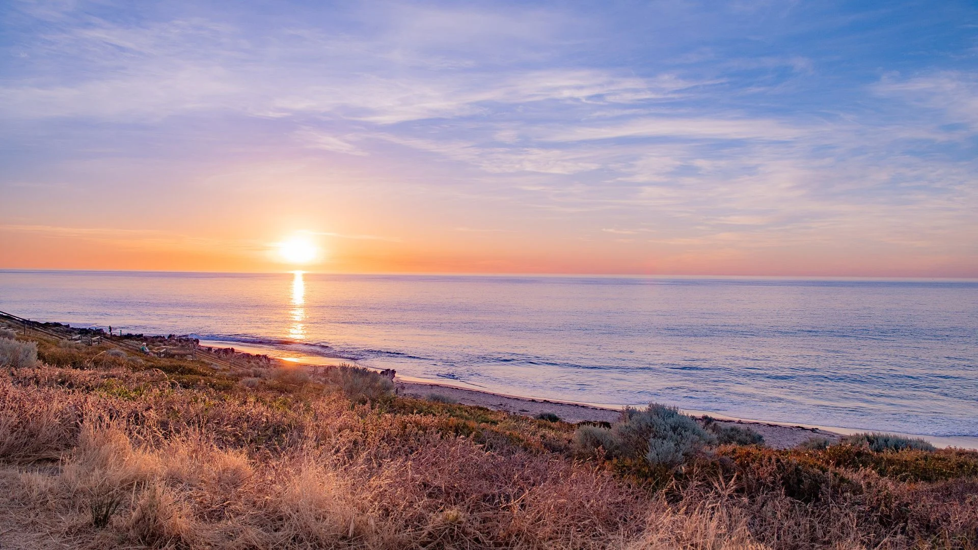 Sunset over a calm ocean with gentle waves, seen from a grassy and bushy shoreline. The sky is partly cloudy, glowing with orange, pink, and purple hues, reflecting on the water’s surface.