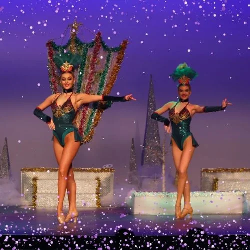Two female dancers in festive green costumes and headpieces perform on stage with holiday decorations, gift boxes, and sparkling tinsel, framed by digital falling snow and a purple background.