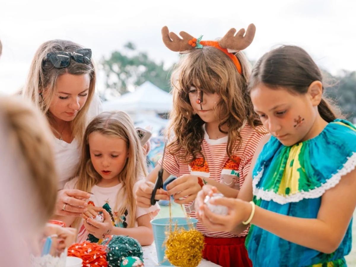 A woman and three girls work on arts and crafts at a table outdoors. One girl wears reindeer antlers, another has a small face painting, and they are all focused on making colorful decorations.