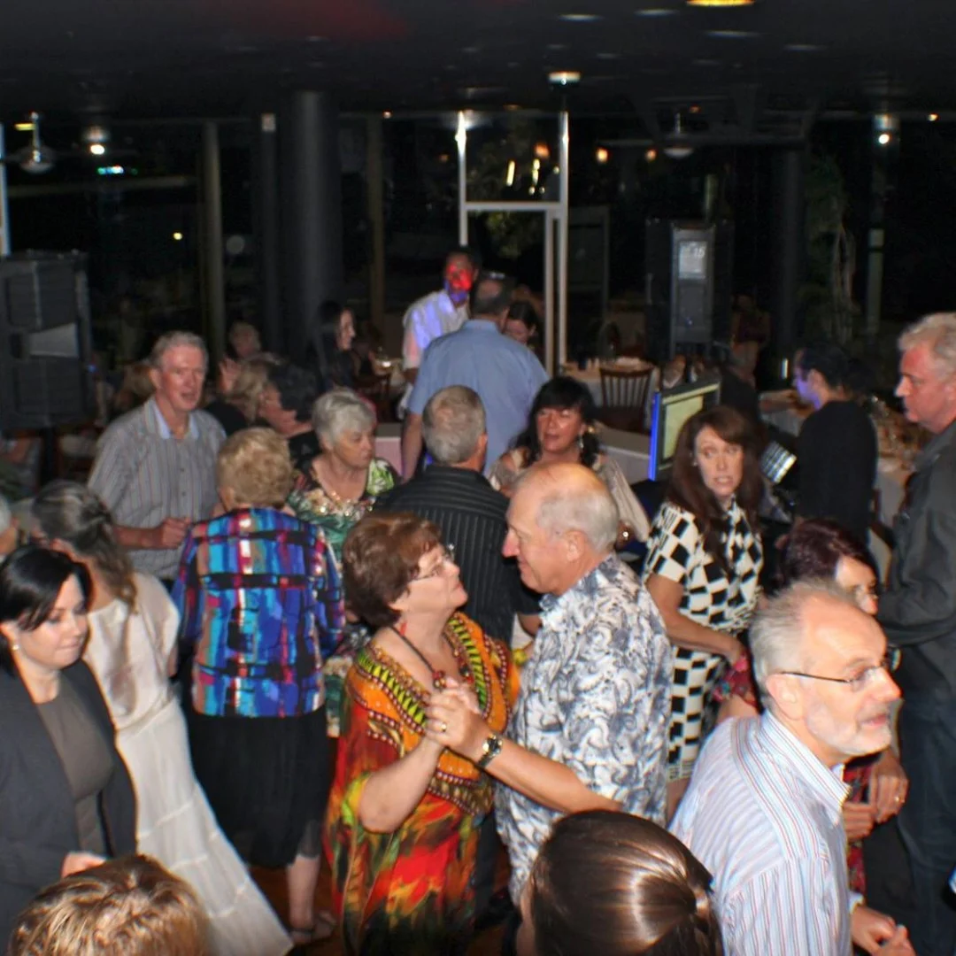 A group of people of various ages dance together in a warmly lit indoor venue. The atmosphere appears festive and lively, with many smiling and engaging in conversation or dancing in pairs.