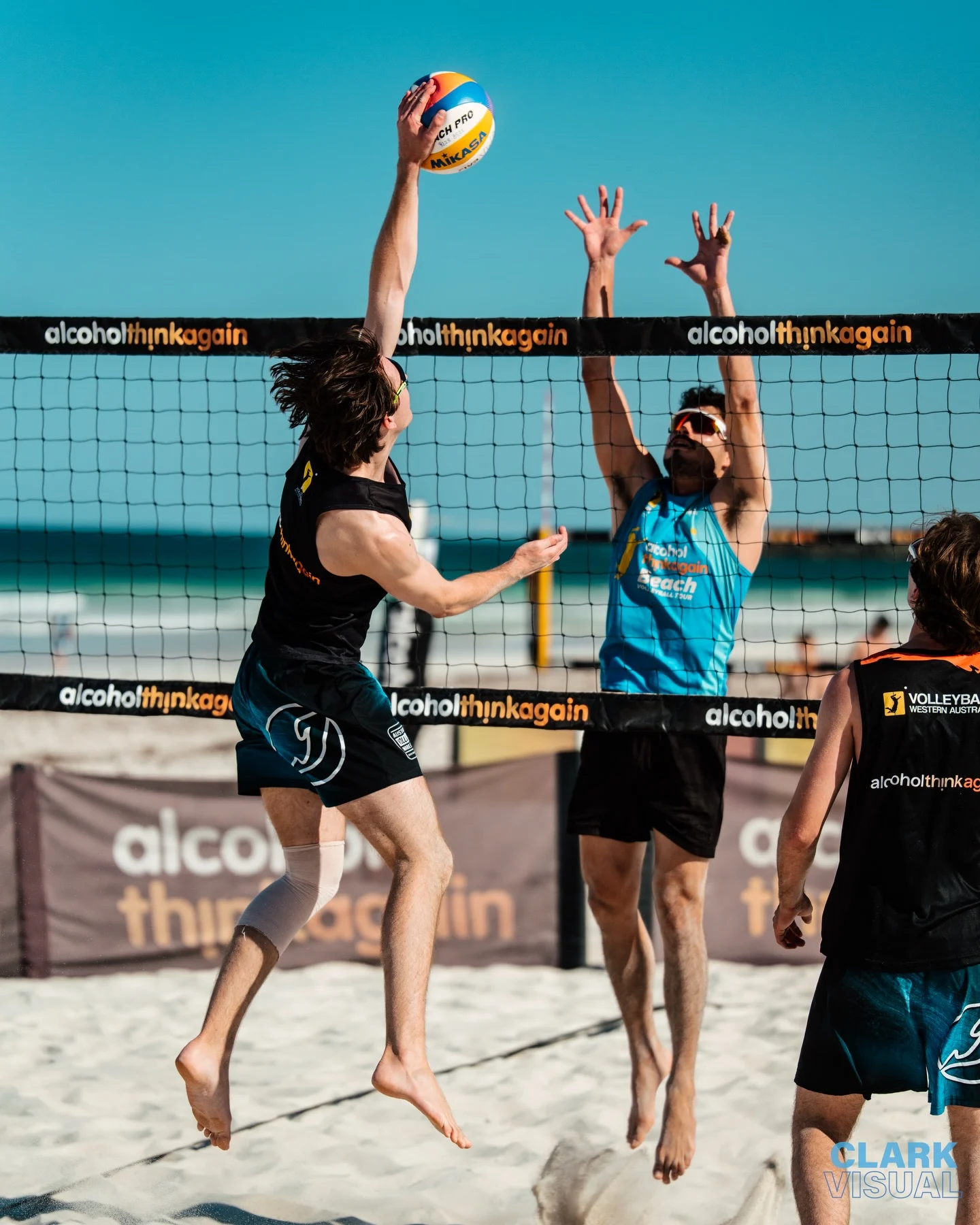 Two men play beach volleyball; one in black jumps to spike the ball while the other in blue jumps to block. Both are on sand near the ocean, under a clear blue sky. The net and branded banners are visible.