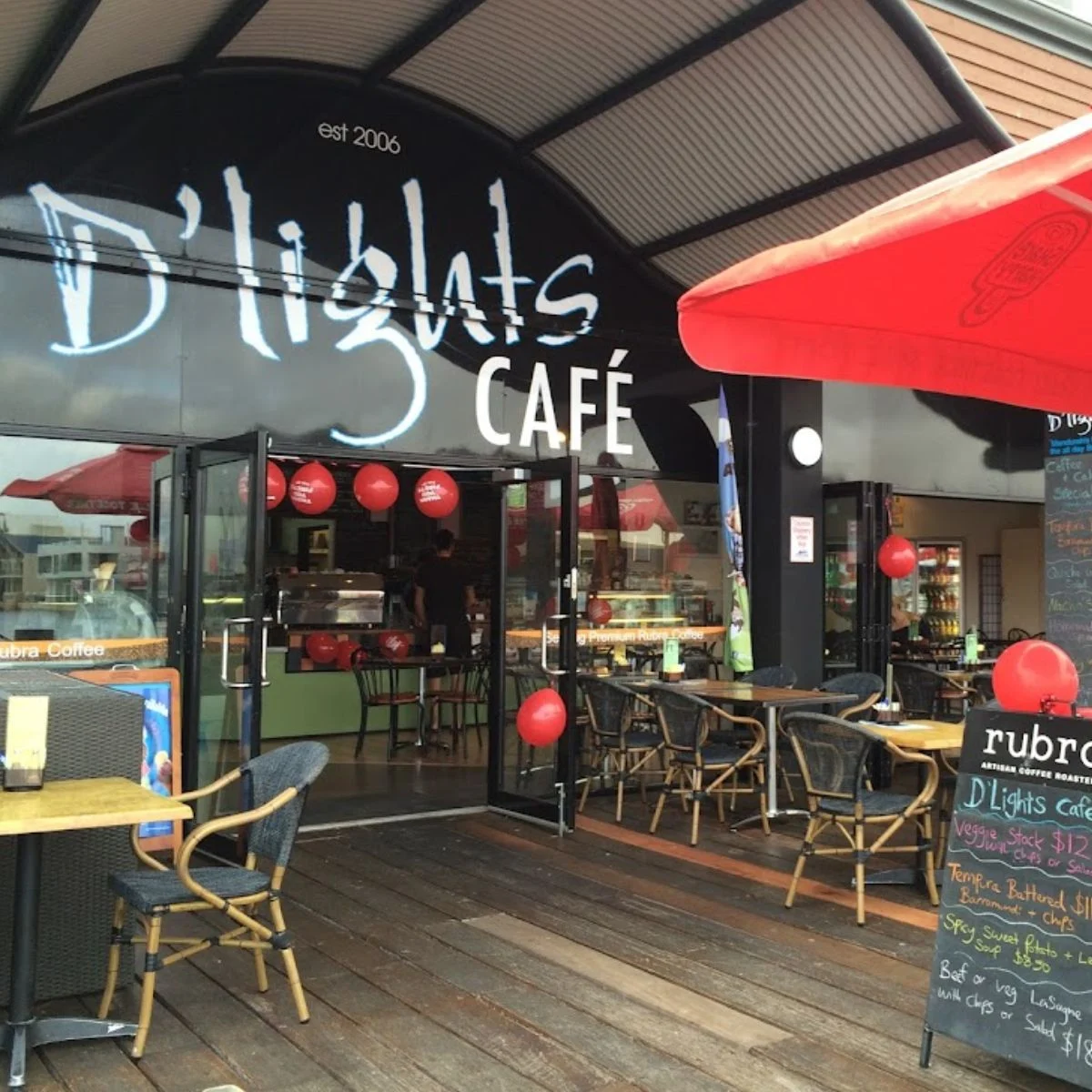 Outdoor view of D’lights Café with open glass doors, red balloons, and several empty tables and chairs on a wooden deck. A chalkboard menu and a large red umbrella are visible in front of the entrance.