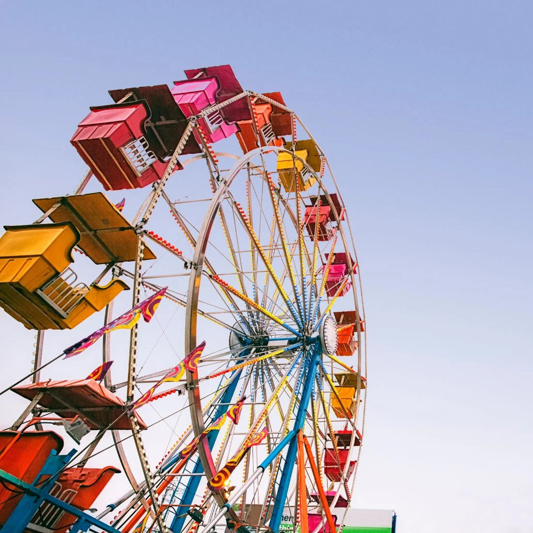 A colorful Ferris wheel with red, yellow, and orange gondolas is seen against a clear blue sky, viewed from a low angle, creating a vibrant and cheerful carnival atmosphere.