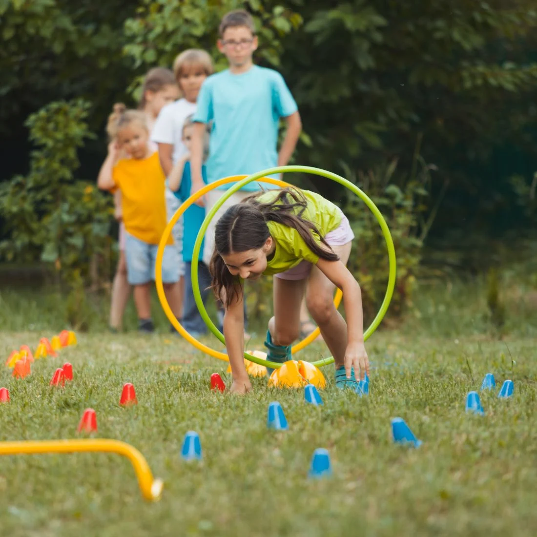 A girl crawls through colorful hoops in an outdoor obstacle course while several children wait in line behind her, with green trees in the background.