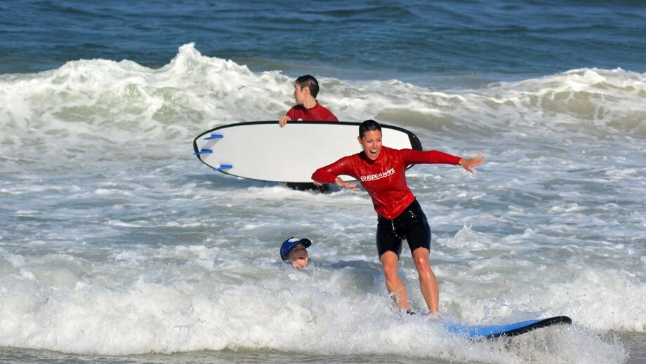 Three people in red shirts surf in shallow ocean waves; one stands on a board, another carries a board, and a child in the water looks on.
