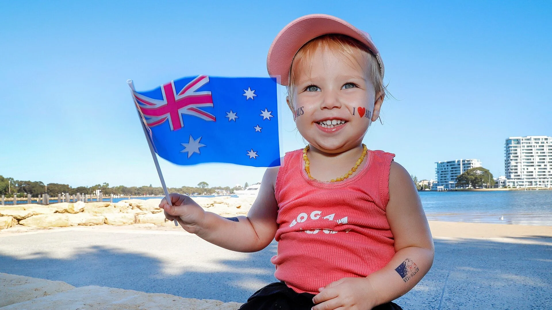 A smiling young child wearing a pink cap and shirt holds an Australian flag. She has face paint of an Australian and French flag on her cheeks, and is sitting outdoors near water with buildings and trees in the background.