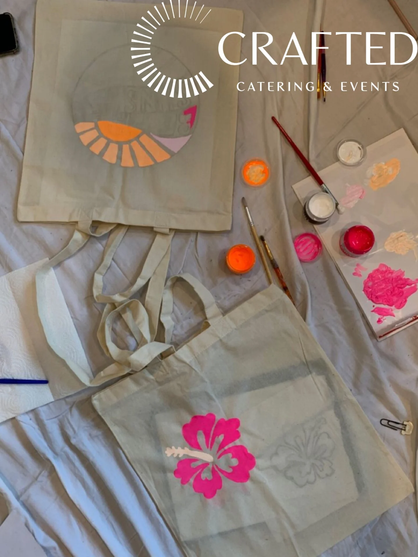 Two canvas tote bags are being painted with colorful flower designs on a light cloth surface. Paintbrushes, paint containers, a palette with pink and orange paint, and a napkin are scattered nearby. CRAFTED Catering & Events logo is visible.
