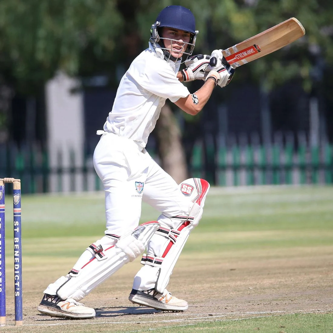 A cricket player in white gear and helmet stands at the crease, holding a bat poised to hit, next to stumps on a grassy field. Trees and a fence are visible in the blurred background.