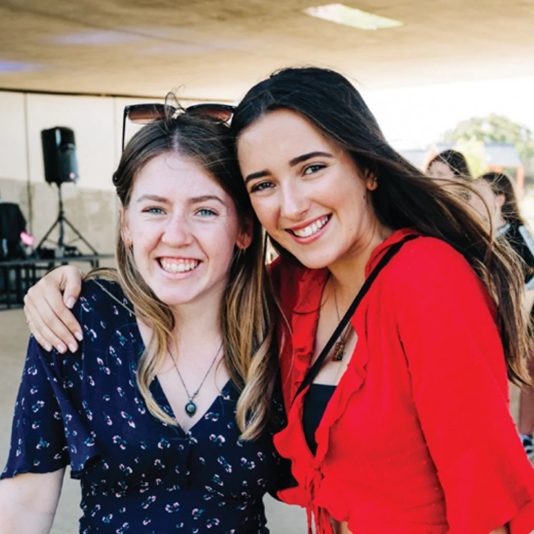 Two young women smile at the camera, standing close with one arm around each other. One wears a navy blue dress with floral print, the other a bright red top. They are outdoors, under a shaded area.