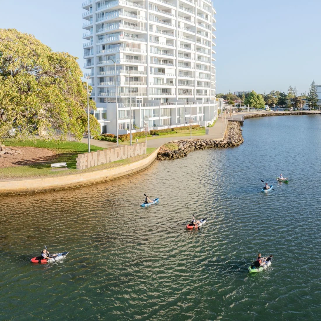 A group of kayakers paddle on a river near a modern white high-rise building and large #MANDURAH sign, with trees and a riverside walkway in the background.
