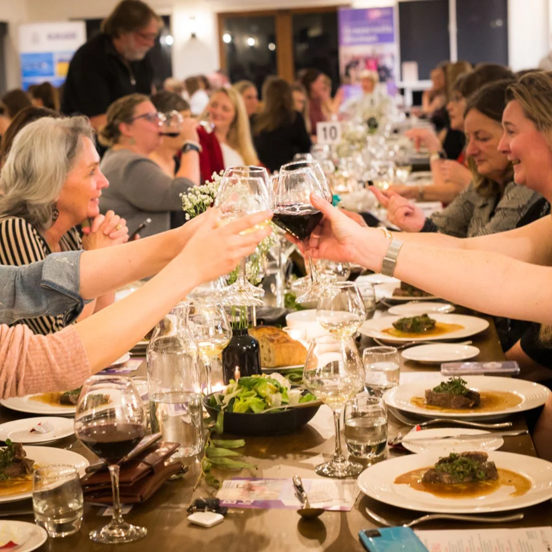 A group of people sit around a long, elegantly set dinner table, raising their wine glasses in a toast. Plates of food, salads, and glasses fill the table, creating a warm, festive atmosphere.