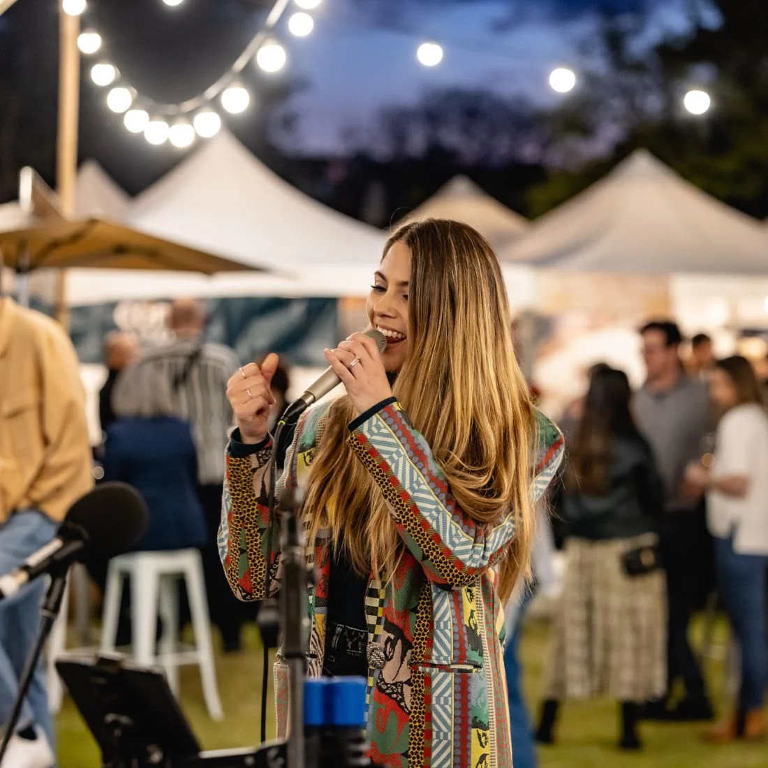 A woman with long hair sings into a microphone at an outdoor event in the evening, surrounded by string lights, tents, and people socializing in the background.