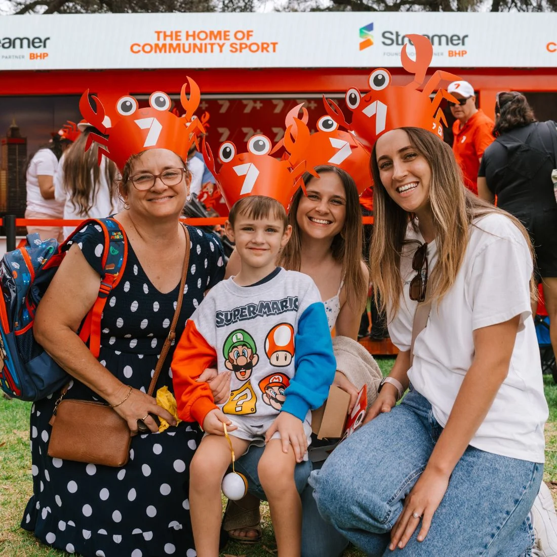 Four people, including a young boy, smile at an outdoor event. They all wear red crab hats. Behind them is a booth with signs about community sports. The group appears happy and festive.