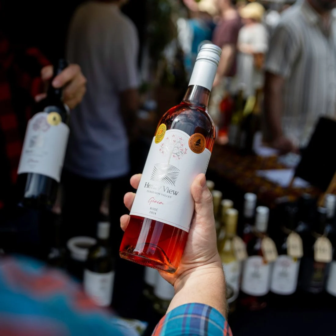 A person holds a bottle of rosé wine at an outdoor market stall, with other wine bottles on the table and people in the background, slightly out of focus.