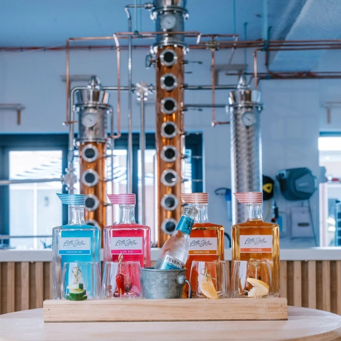 Four colorful bottles of gin with different labels sit on a wooden tray with garnished glasses, a bottle of tonic water in an ice bucket, and a distillery setup with copper stills in the background.