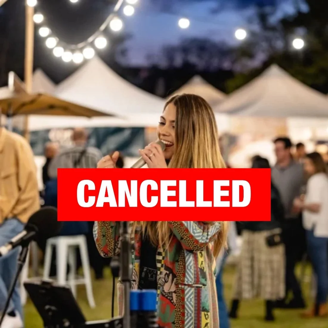 A woman sings into a microphone at an outdoor event with string lights and tents. A red CANCELLED banner is overlaid across the image, partially obscuring her. People and booths are visible in the background.