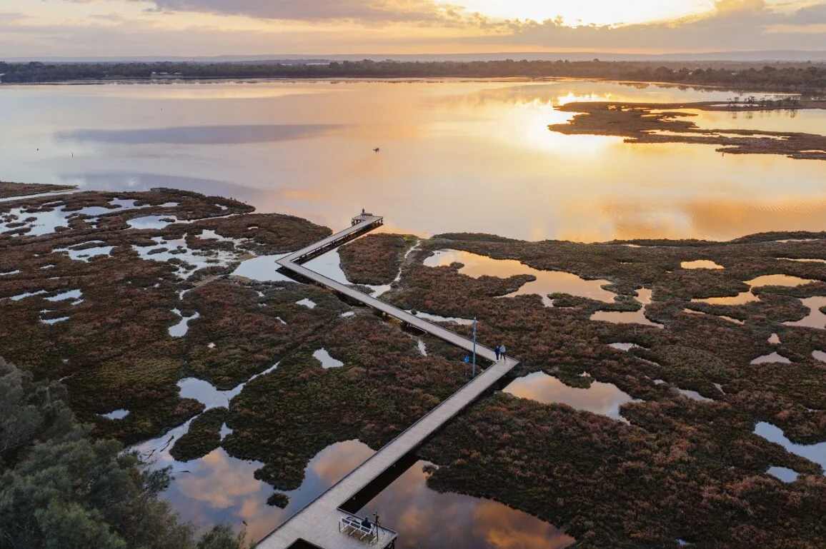 A winding boardwalk stretches over wetlands at sunset, with calm water reflecting the orange and yellow hues of the sky. Small patches of vegetation dot the landscape, creating a tranquil, scenic view.