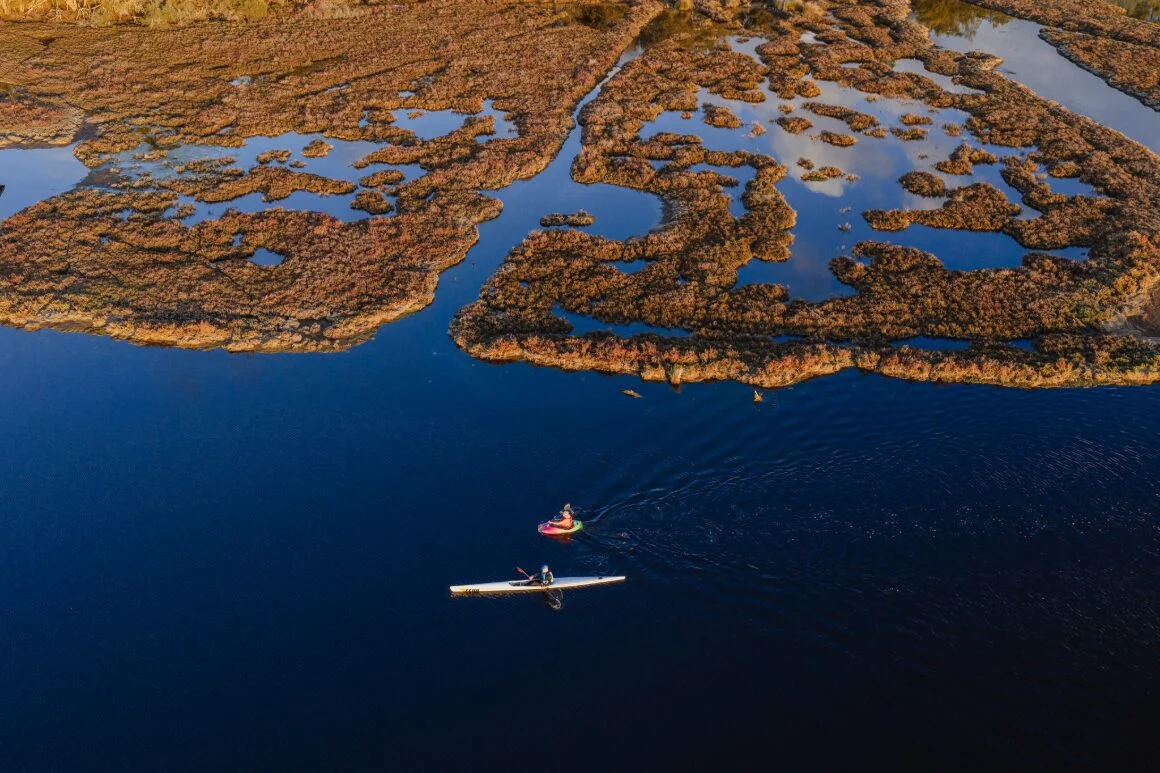 Aerial view of two people kayaking in deep blue water, surrounded by patches of brown marshland and reflecting sky, creating a striking natural landscape.