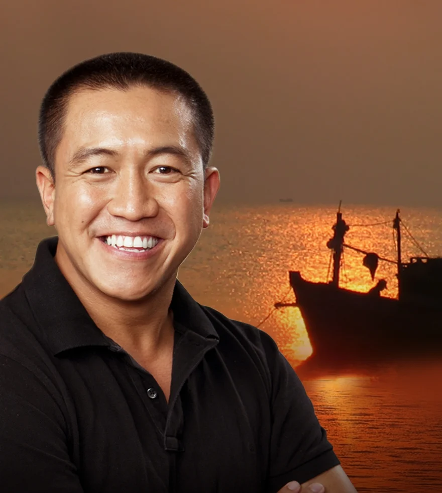 A smiling man in a black shirt stands in the foreground, with a sunset over the ocean and the silhouette of a boat in the background.