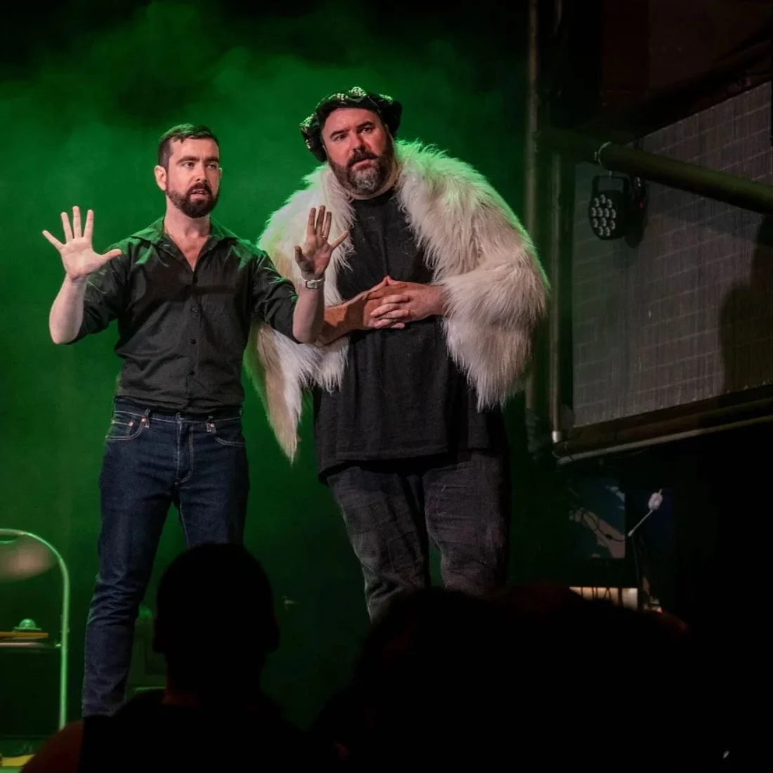 Two performers on stage, one in a black shirt and jeans, the other in a black t-shirt and jeans wearing a white fur stole and a black cap.