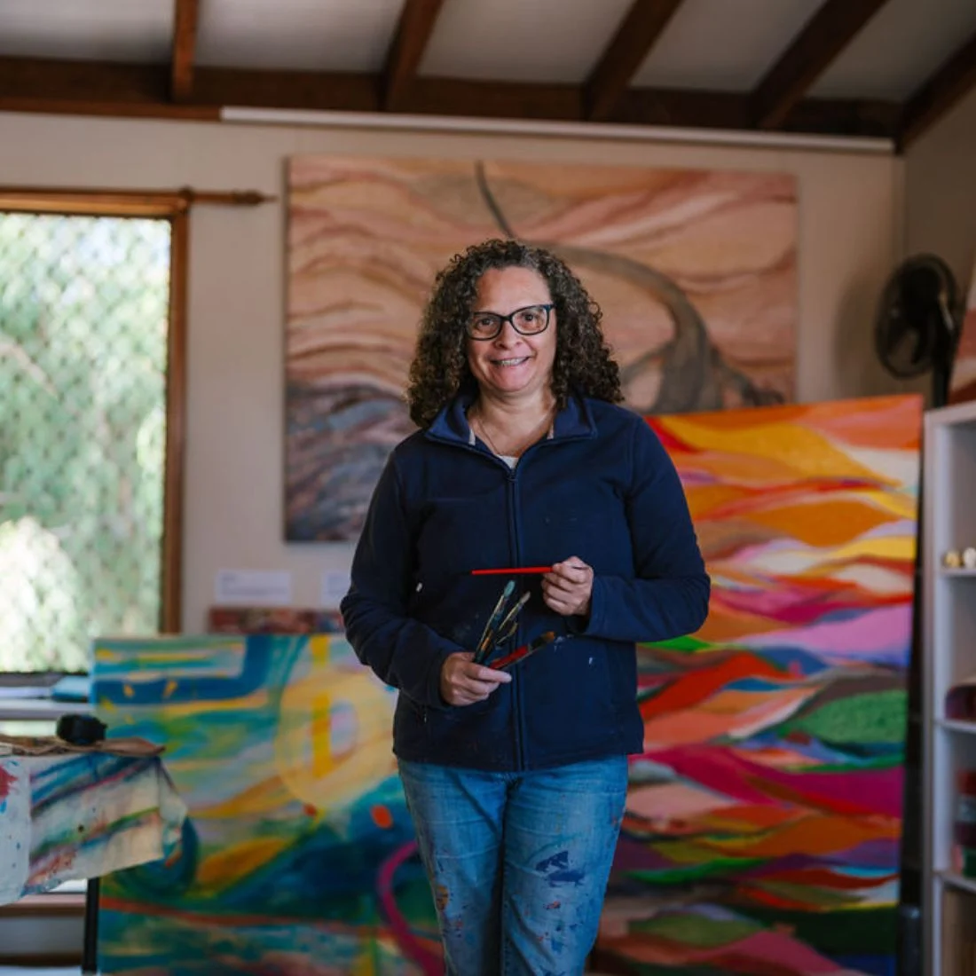 A woman with curly hair and glasses, wearing a navy jacket and paint-splattered jeans, stands smiling in an art studio holding paintbrushes, surrounded by colorful abstract paintings.