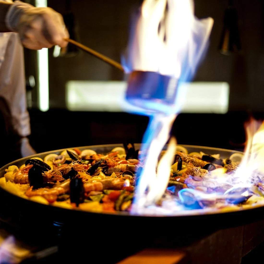 A chef uses a ladle to flambé a large pan of seafood paella, with vibrant flames rising above the colorful dish of shrimp, mussels, and rice.